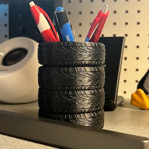 May include: A black pen holder made of stacked tire replicas, holding three pens. The pens are red, white, and blue. The pen holder sits on a desk with other office items in the background.