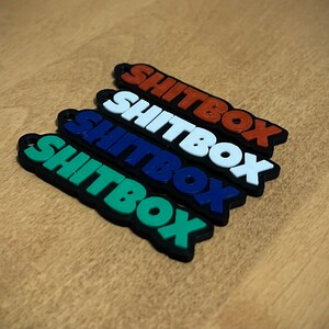 Pode incluir: Quatro chaveiros com a palavra "SHITBOX" em diferentes cores: verde, azul, branco e laranja. Os chaveiros são feitos de um material emborrachado.