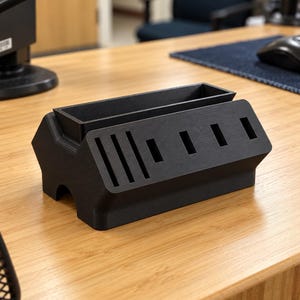 May include: A matte black desk organiser featuring a rectangular storage compartment and a front-facing panel with various vertical and horizontal slots designed for holding small items, constructed with a textured, 3D-printed finish.