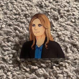 May include: A clear acrylic keychain featuring a cartoon illustration of a woman with long blonde hair, wearing a black blazer over a blue shirt.