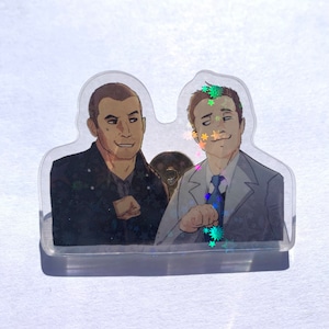 Puede incluir: Dos hombres están representados en un estilo de dibujos animados, uno con una chaqueta de cuero negra y el otro con un traje gris. El fondo es una superficie brillante e iridiscente.