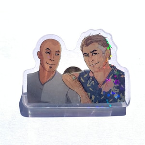 May include: A clear acrylic stand with two cartoon figures. The figure on the left is a man with brown skin, short black hair, and a gray shirt. The figure on the right is a man with light skin, short brown hair, and a blue and white patterned shirt.