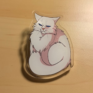 May include: A white cat with blue eyes sits with its tail curled around its body. The cat has a grumpy expression on its face. The image is printed on a clear acrylic keychain.