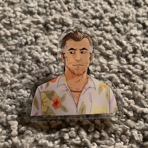 May include: A man with a beard and short gray hair is wearing a white button-down shirt with a floral pattern. The shirt has yellow, orange, and purple flowers.
