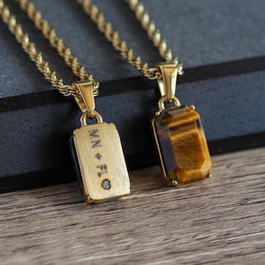 May include: Two gold-colored necklaces with rectangular pendants. One pendant has the engraved text "MN + FLO" and the other features a tiger's eye stone. The necklaces are displayed on a wooden surface.