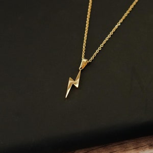 Puede incluir: Un collar de cadena dorada con un pequeño colgante de rayo. El collar está sobre una superficie negra.