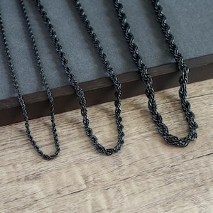 Puede incluir: Tres cadenas de cuerda negras de diferentes grosores dispuestas en fila. Cada cadena presenta un diseño de eslabones metálicos trenzados con un acabado mate oscuro. Las cadenas están colocadas sobre un expositor rectangular gris oscuro con el texto MEN JEWELS.
