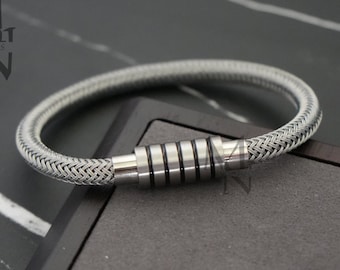 May include: A silver-toned braided cord bracelet featuring a cylindrical metal clasp with five distinct segments separated by thin black rings. The logo text MEN JEWELS is visible in the upper left corner of the frame.