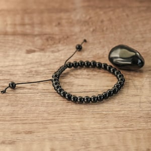 May include: A black beaded bracelet with a black stone charm. The bracelet is made of small, round beads and has a simple, adjustable cord closure. The stone charm is smooth and polished.