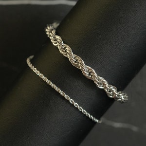 May include: Two silver rope chain bracelets on a black display stand. The thicker chain is on top of the thinner chain.