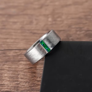 Könnte beinhalten: Ein silberfarbener Ring mit gebürsteter Oberfläche. Ein vertikaler Streifen aus kleinen, leuchtend grünen Steinen ist in das Band des Rings eingesetzt. Der Ring wird auf einer dunklen Oberfläche vor einem Holzgrund gezeigt. Der Text "MEN JEWELS" ist sichtbar.