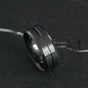 Könnte beinhalten: Ein schwarzer Ring mit gerilltem Design. Der Ring hat eine matte Oberfläche und einen schlichten, modernen Stil. Der Ring ist wahrscheinlich aus Metall und wird auf einer dunklen Oberfläche präsentiert.