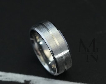 Peut inclure: Une bague en métal argenté avec une finition brossée mate et une rainure centrale polie. La bague présente un design à bords biseautés. Le texte "MG MEN JEWELS" est visible dans le coin supérieur gauche et à droite de l'image.