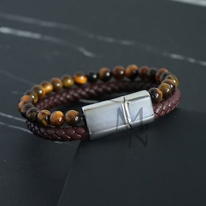 Puede incluir: Un brazalete de cuero marrón con cierre magnético y dos filas de cuentas de ojo de tigre.