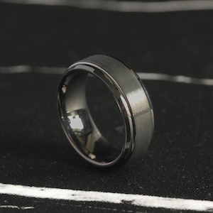 Puede incluir: Un anillo negro y plateado con un borde biselado. El anillo está hecho de un metal duradero y tiene un diseño elegante y moderno.