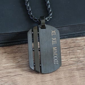 Puede incluir: Un collar con colgante de chapa de identificación negra con una cadena a juego. La chapa tiene cortes verticales y números romanos grabados. El collar descansa sobre una superficie oscura y una mesa de madera.