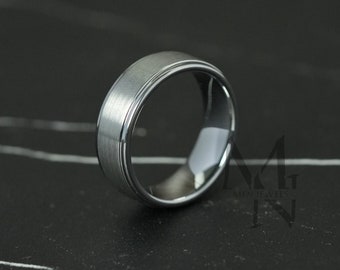 Peut inclure: Une bague en métal argenté avec une finition mate brossée sur la bande centrale et des bords polis et réfléchissants. La bague présente un détail rainuré subtil le long de la circonférence, créant un aspect superposé. Le logo MG MEN JEWELS est visible.