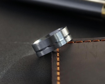 Peut inclure: Une bague en métal gris brossé présentant une rainure horizontale unique faisant le tour du centre de l'anneau. La bague a un profil plat avec une finition mate, mettant en valeur un design épuré qui souligne la texture métallique du matériau.