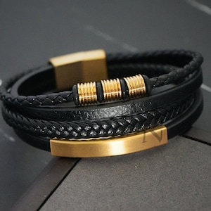 Op de afbeelding: Een zwarte leren armband met gouden accenten. De armband heeft een gevlochten leren band en een gouden balk met een kleine gouden kraal.