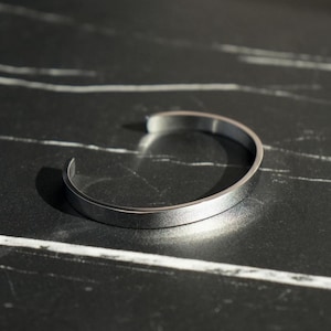 May include: A silver bangle bracelet with a smooth, polished finish. The bracelet is open and has a simple, minimalist design.