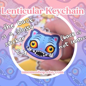 May include: A lenticular keychain featuring a blue cat-like creature with large yellow eyes and sharp teeth. The keychain has a glossy sticker back. The background is a gradient of pink and blue with repeating cat-like creature faces.