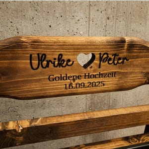 Puede incluir: Banco de madera con texto grabado. El texto incluye los nombres "Ulrike" y "Peter" separados por un coraz&oacute;n, y las palabras "Goldene Hochzeit 16.09.2025". La madera tiene un tono marr&oacute;n c&aacute;lido.