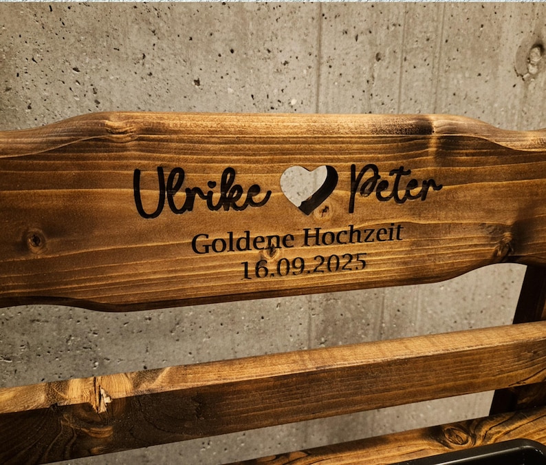 Puede incluir: Banco de madera con texto grabado. El texto incluye los nombres "Ulrike" y "Peter" separados por un coraz&oacute;n, y las palabras "Goldene Hochzeit 16.09.2025". La madera tiene un tono marr&oacute;n c&aacute;lido.