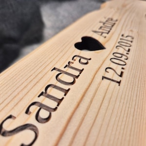 Puede incluir: Un letrero de madera con los nombres "Sandra" y "Andre" grabados, un coraz&oacute;n negro y la fecha "12.09.2015". El letrero es de madera clara.
