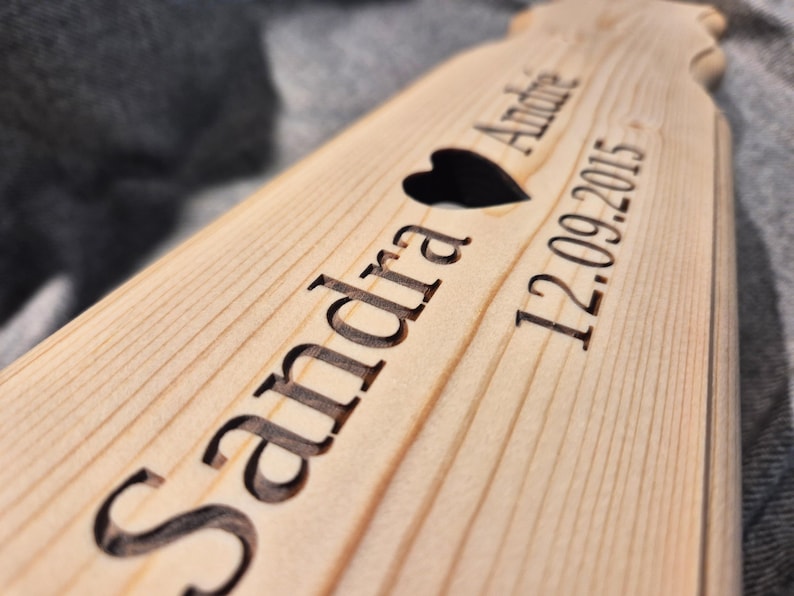 Puede incluir: Un letrero de madera con los nombres "Sandra" y "Andre" grabados, un coraz&oacute;n negro y la fecha "12.09.2015". El letrero es de madera clara.