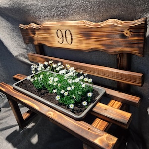 Puede incluir: Un banco de madera r&uacute;stico con una jardinera llena de plantas verdes y flores blancas. El respaldo del banco presenta el n&uacute;mero "90" grabado en la madera. El banco es de madera te&ntilde;ida oscura.