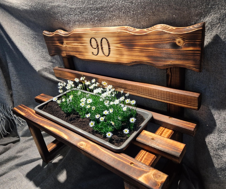Puede incluir: Un banco de madera r&uacute;stico con una jardinera llena de plantas verdes y flores blancas. El respaldo del banco presenta el n&uacute;mero "90" grabado en la madera. El banco es de madera te&ntilde;ida oscura.