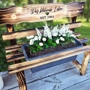 Puede incluir: Banco de madera con una jardinera llena de flores blancas y vegetaci&oacute;n. El respaldo del banco tiene el texto "Das bl&uuml;hende Leben" y "SEIT 1964". El banco es de madera con un acabado oscuro y quemado.
