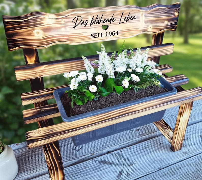 Puede incluir: Banco de madera con una jardinera llena de flores blancas y vegetaci&oacute;n. El respaldo del banco tiene el texto "Das bl&uuml;hende Leben" y "SEIT 1964". El banco es de madera con un acabado oscuro y quemado.