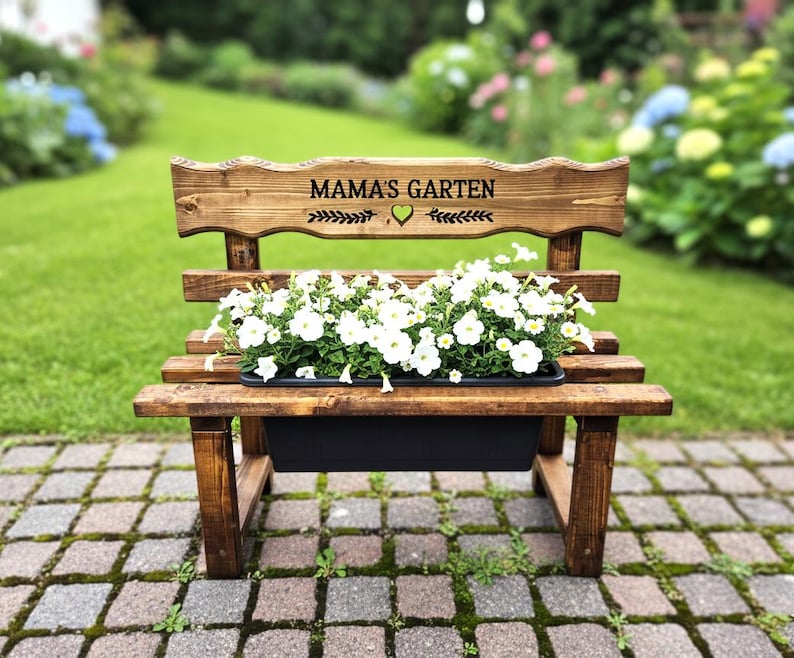 Könnte beinhalten: Gartenbank aus Holz mit dem Text "MAMA'S GARTEN" auf der Rückenlehne. Die Bank hat einen schwarzen Pflanzkasten mit weißen Blumen. Die Bank ist aus gebeiztem Holz gefertigt und steht auf einer Backstein-Terrasse in einem Garten.