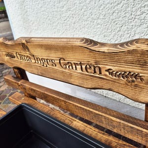 Puede incluir: Banco de jard&iacute;n de madera con el texto grabado "Oma Inges Garten" y un dise&ntilde;o de espiga de trigo. El banco tiene un tinte marr&oacute;n oscuro y una jardinera negra. El banco es de madera.