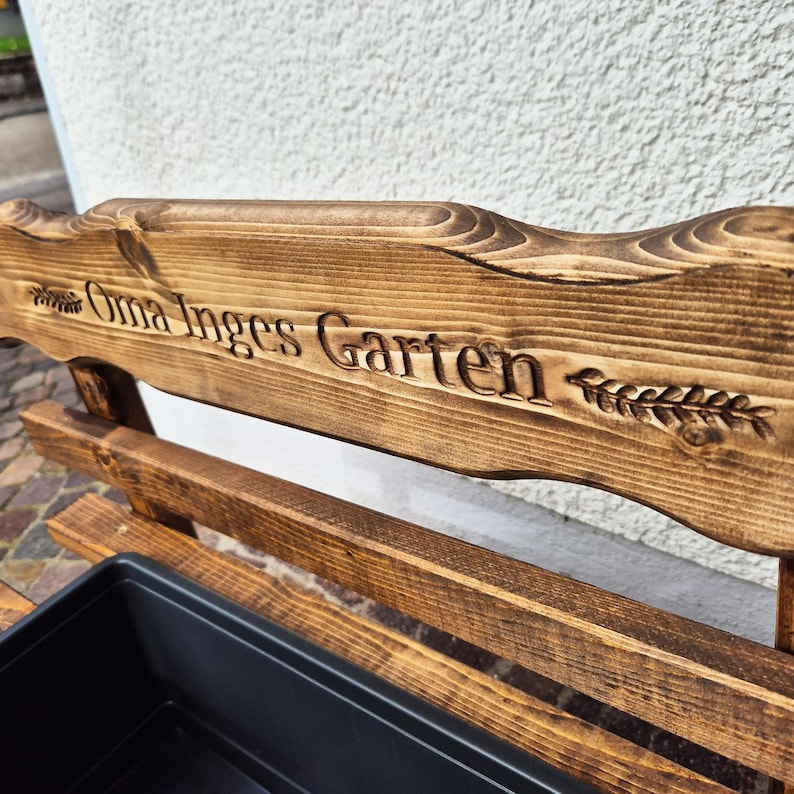 Puede incluir: Banco de jard&iacute;n de madera con el texto grabado "Oma Inges Garten" y un dise&ntilde;o de espiga de trigo. El banco tiene un tinte marr&oacute;n oscuro y una jardinera negra. El banco es de madera.