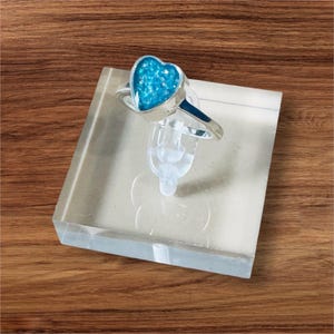 Peut inclure: Une bague en argent avec un centre en forme de cœur bleu clair. La bague est présentée sur un bloc acrylique transparent, sur un fond en bois. Le centre en forme de cœur a un aspect scintillant.