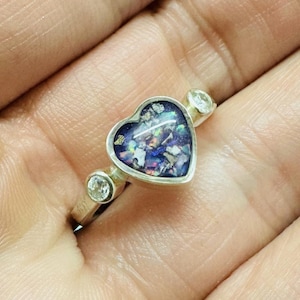 May include: A silver ring featuring a heart-shaped setting filled with shimmering, multi-coloured fragments. The ring includes two clear, round accent stones. The heart is framed in silver.