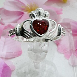 May include: A silver Claddagh ring with a red heart-shaped stone. The ring is on a white display stand.