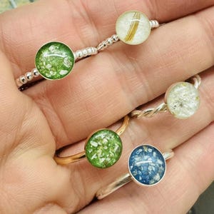 May include: Five silver rings with different coloured resin insets. The resin insets are filled with various materials, including green, blue, and white.