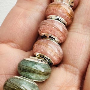 Peut inclure: Six perles de verre avec des cheveux intégrés et des accents argentés. Les perles sont dans des tons de rose et de vert. Les perles sont gravées des mots "Always", "With", "She", et "Love".