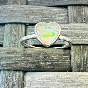 Peut inclure: Une bague en argent avec une pierre précieuse en opale en forme de cœur. L'opale est irisée avec des nuances de blanc, de vert et de bleu.