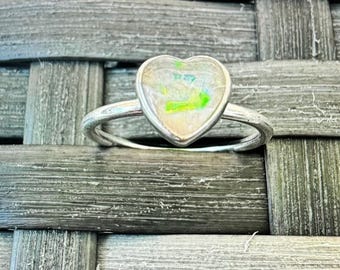 May include: A silver ring with a heart-shaped opal gemstone. The opal is iridescent with shades of white, green, and blue.