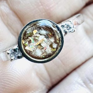 May include: A silver ring with a round, clear center filled with a mix of brown, white, and iridescent materials. Two small, clear stones are set on either side of the central design.