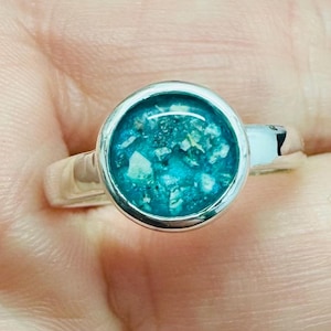 May include: A silver ring with a round, turquoise-colored center. The center has a textured appearance with flecks of white and other colors. The ring is set in a polished silver band.