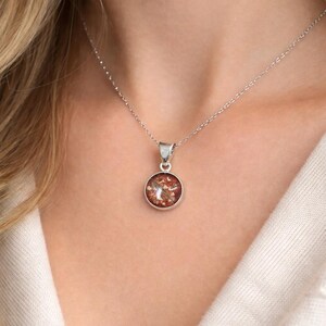 May include: A silver necklace with a round pendant. The pendant features a reddish-brown design with a silver bezel. The necklace is worn on a person wearing a white sweater.