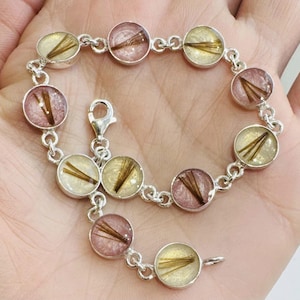 Peut inclure: Un bracelet en argent avec des breloques rondes contenant des mèches de cheveux enfermées dans de la résine transparente. Les breloques alternent les teintes rose et blanche.