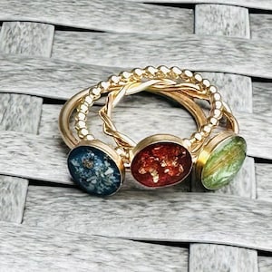 Könnte beinhalten: Drei goldene Ringe mit farbigen, oval geformten Mittelsteinen. Die Steine sind blau, rot und grün. Die Ringe liegen auf einer grauen, gewebten Oberfläche.