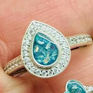 Peut inclure: Une bague en argent avec un sertissage en forme de larme présentant une pierre bleue et verte avec des paillettes dorées. Le sertissage est entouré d'un halo de petites pierres claires.