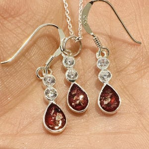 Könnte beinhalten: Ein silbernes Halsketten- und Ohrring-Set mit tropfenförmigen Anhängern mit einem rot-weiß gesprenkelten Design. Jedes Stück hat drei klare, runde Akzente und eine silberne Kette oder Haken.